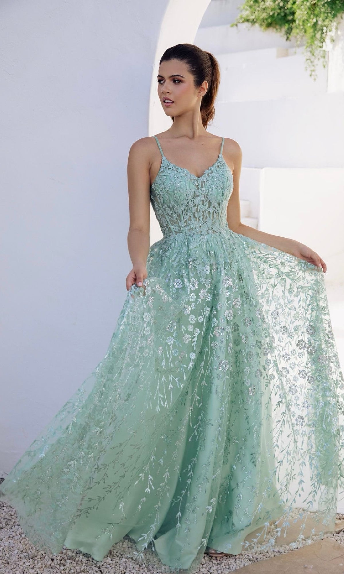 A woman wears the Eureka 9908 Long Glitter A-Line Prom Dress, featuring a light green hue and sheer floral overlay, as she stands outdoors by a white wall, gracefully holding out her gown.